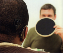View from the back of the patient. A brown hearing aid is visible on one ear. A clinician holding a round black mat to hide his mouth is seen sitting in front of the patient.