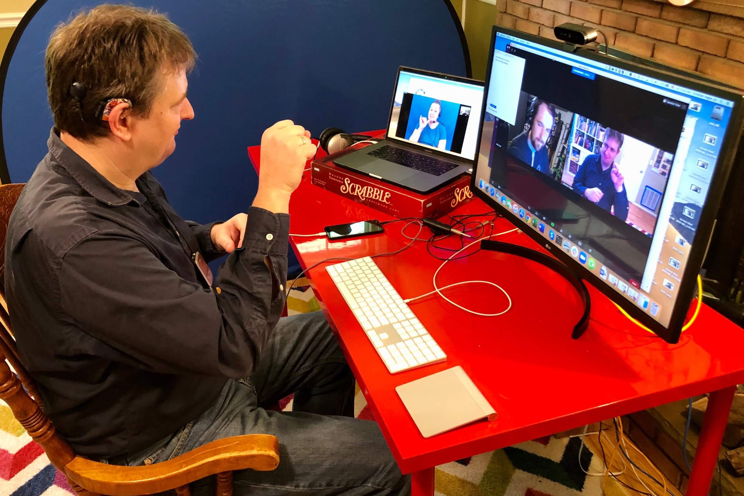 Man signing during a teleconference. He is sitting in front of a laptop and an external monitor in a home environment. A video relay service interpreter is on the laptop screen, while the videoconference is on the external monitor.