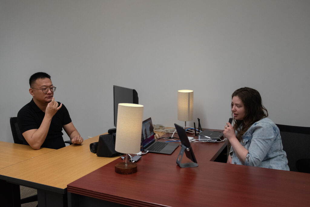 An Asian man and a White woman are sitting at desks across from each other. The man is signing "Light" in American Sign Language, and the woman is speaking into a microphone while looking at a laptop. The man has short black hair and wears a black T-shirt. The woman has long brown hair and wears a light blue button-up shirt. There is a laptop, two lamps, and cords on both desks.
