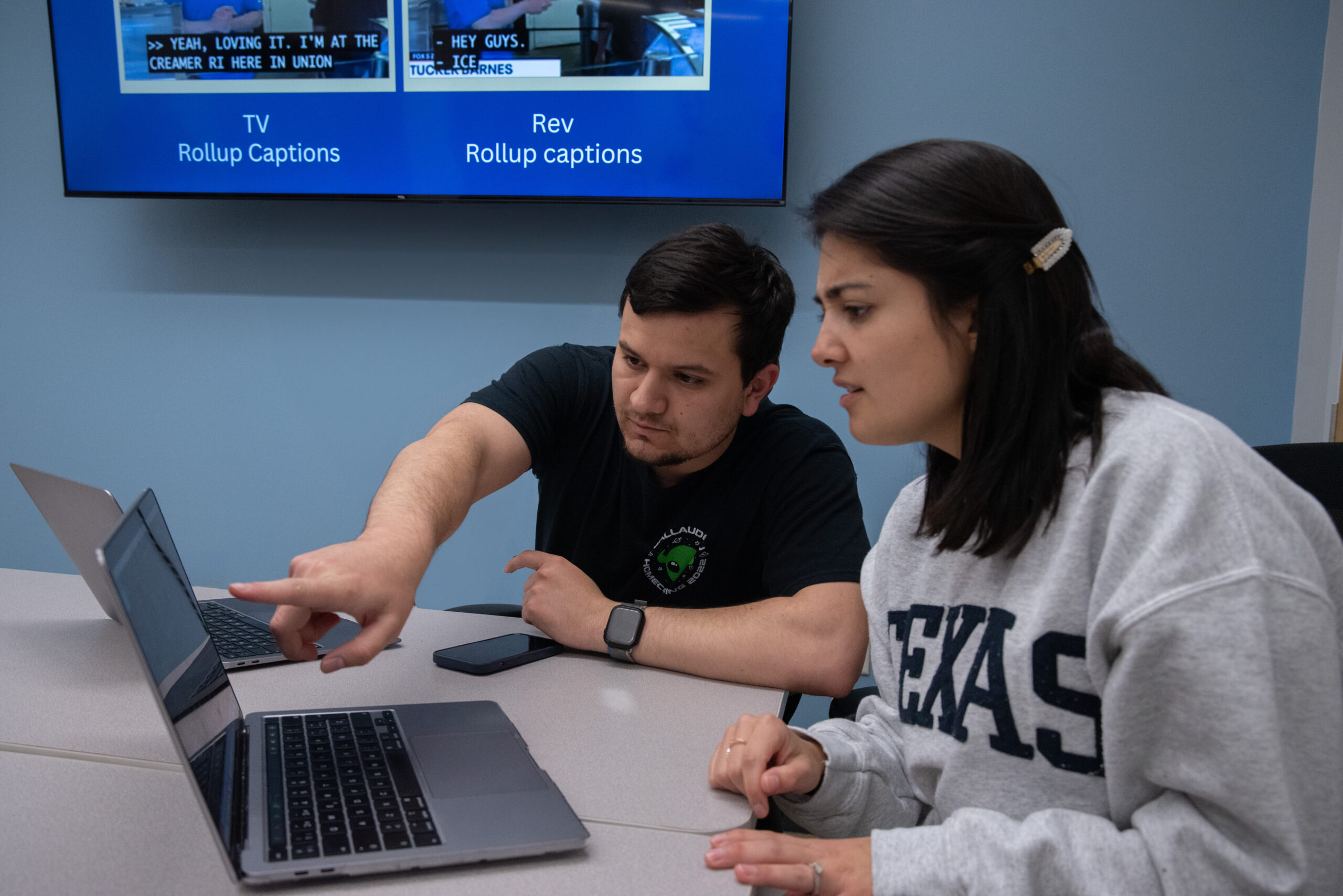A White Hispanic man points at a laptop screen while signing next to a Hispanic woman. They both look focused on reviewing captioning styles during a meeting. The man has dark brown hair and wears a black shirt with a green alien on the left side. The woman has short, straight black hair and wears a gray sweatshirt with big letters reading “Texas.” The big-screen TV behind them shows a comparison between two types of captions: TV and REV rollup.